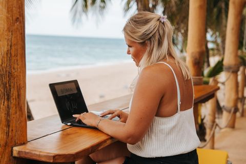 Manon working on a laptop at a wooden beach bar, the sea behind her