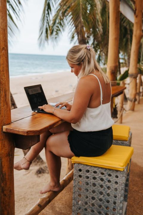 Manon working on her laptop at a beach bar, bare feet resting on the wooden rail