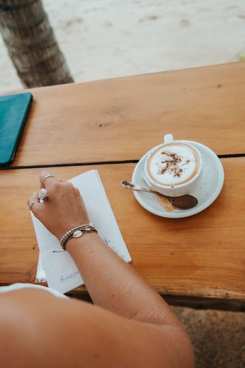 A hand writing in a notebook next to a cappuccino on a wooden table