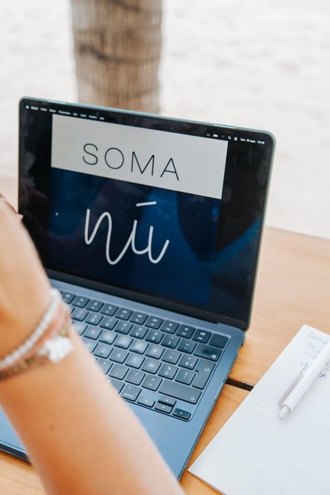 Laptop on a wooden table showing the SOMANÚ logo, a notebook and pen beside it