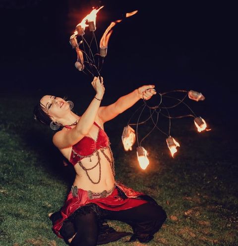 A fire performer kneeling on the grass at night, holding flaming fans