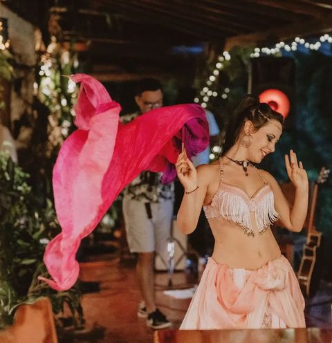 A dancer swirling a pink silk veil at an evening event among plants and string lights
