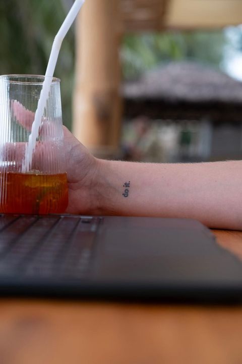 Close-up of a wrist with a small “do it.” tattoo, next to an iced tea and a laptop