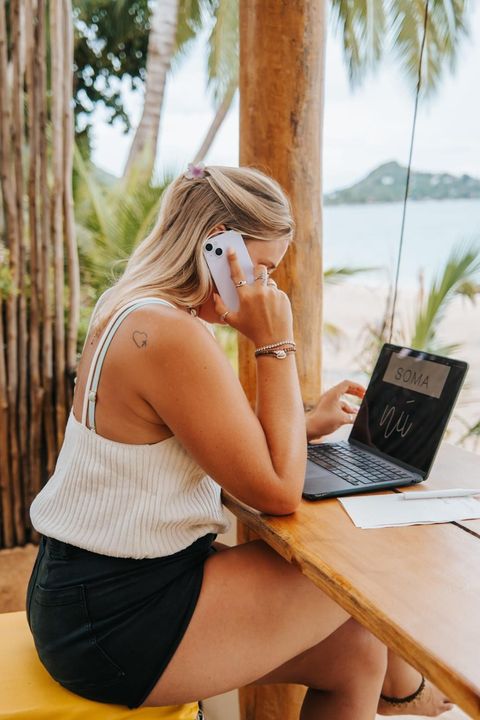 Manon on the phone at a beach bar, laptop open in front of her, the bay behind